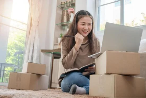 asian woman celebrating over laptop while surrounded by cardboard boxes